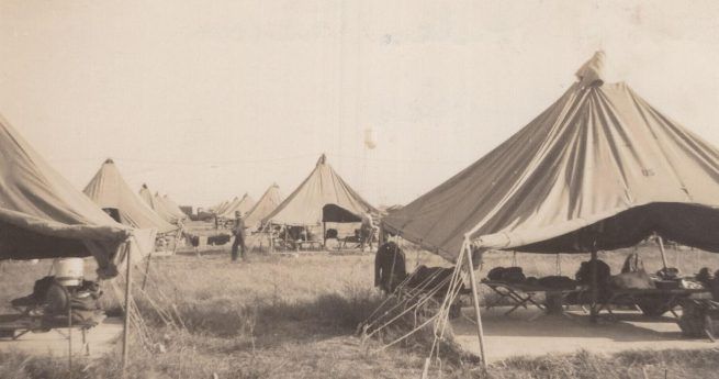 A black-and-white photograph of a military camp at Camp Hulen, Texas, featuring several large canvas tents arranged in rows on a grassy field. The tents are pyramid-shaped with open sides, revealing cots and personal belongings of the soldiers inside. A few soldiers can be seen walking around the camp, with some tents in the background, creating a sense of daily life and routine at the camp.