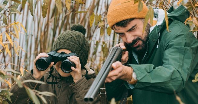 A father and son stand hidden among tall reeds while duck hunting. The father, dressed in a green raincoat and orange beanie, is aiming a shotgun, while the son, wearing a green knit hat and brown jacket, uses binoculars to scan the surroundings.