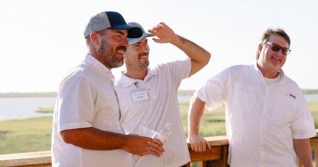 Three men in light-colored shirts stand together on a wooden deck, enjoying a sunny day by the coast.