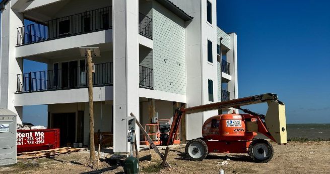 A large, partially constructed building with multiple balconies and a lift machine parked nearby, along with a portable restroom and construction materials around the site.