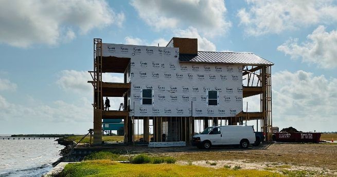 A waterfront house in the early stages of construction, with wooden framing and a few workers on site, along with a van and construction materials nearby.