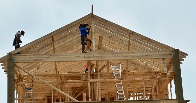 how to buy land and build-men building house-2 Three construction workers building the wooden frame of a house, with one worker on the roof using tools to secure the structure, surrounded by ladders and scaffolding.