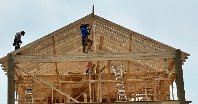 Three construction workers building the wooden frame of a house, with one worker on the roof using tools to secure the structure, surrounded by ladders and scaffolding.