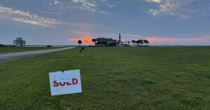 Sunset over a coastal property with a large green lawn, a road, and two houses in the background. In the foreground, a 'Sold' sign stands in the grass, marking the land as sold.