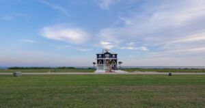 A blue two-story coastal home with white trim sits isolated on a large green lawn, facing a calm body of water under a clear sky with scattered clouds.