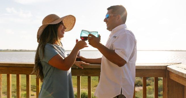 A smiling couple toasts with wine glasses on a wooden deck overlooking a peaceful body of water. The woman, wearing a sunhat and a blue shirt, holds a bottle of wine, while the man, dressed in a white button-up shirt and sunglasses, faces her with his glass raised.