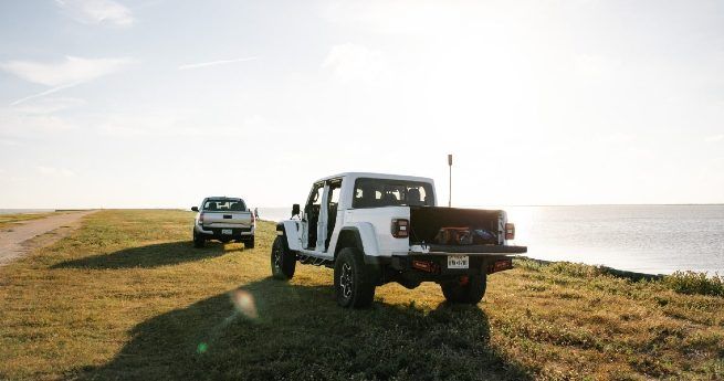 Two vehicles parked on a grassy shore near a body of water under a bright sun.