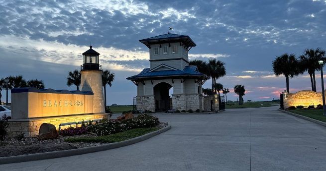 An illuminated entrance to a beachside community at dusk, featuring a sign with the word 'Beachside' and a small lighthouse structure beside it. A stone pavilion with a blue roof sits in the background, flanked by palm trees.