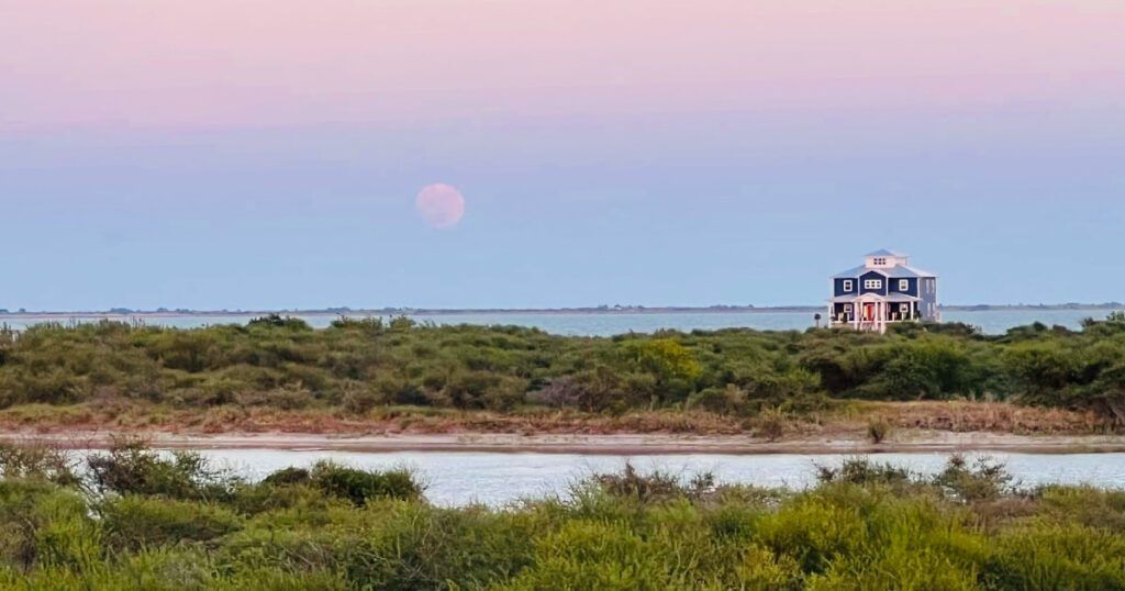 A scenic view of a blue house on stilts surrounded by greenery and water, with a pastel-colored sky at dusk. A large, pinkish full moon is visible near the horizon.