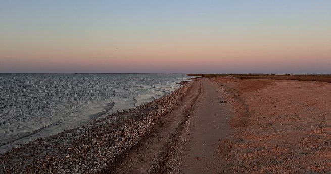 how to finance land in texas-shoreline-4 A quiet shoreline with gentle waves lapping against the sand and pebbles. The beach stretches into the distance under a soft pastel sky at dusk, with a mix of pink and blue hues.