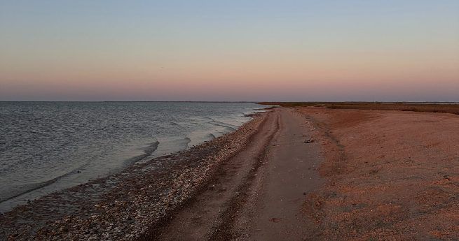 A quiet shoreline with gentle waves lapping against the sand and pebbles. The beach stretches into the distance under a soft pastel sky at dusk, with a mix of pink and blue hues.