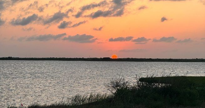 A tranquil sunset over a body of water with an orange sun partially dipping below the horizon.