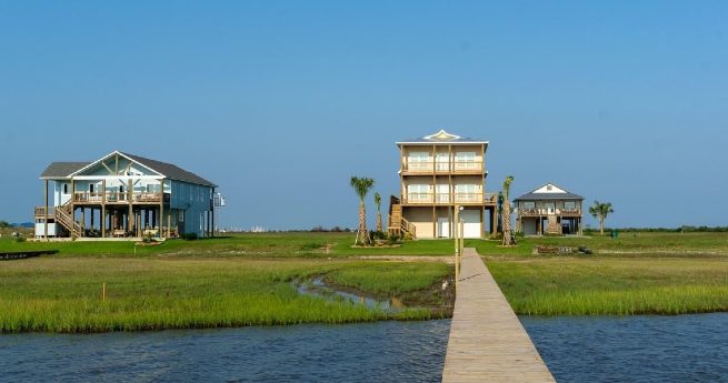 how to finance land in texas-two beachside houses-2 Three beachside homes situated in a grassy waterfront area. A wooden walkway extends from the water toward the center house.