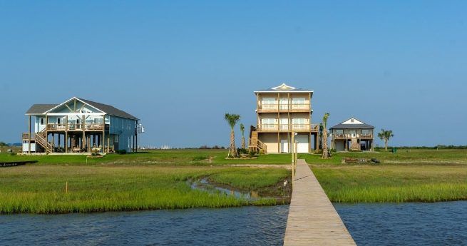 Three beachside homes situated in a grassy waterfront area. A wooden walkway extends from the water toward the center house.