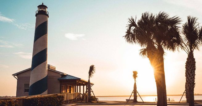 A lighthouse with a black and white spiral pattern stands beside a small building at sunset, with palm trees framing the scene against a bright sky near the water.