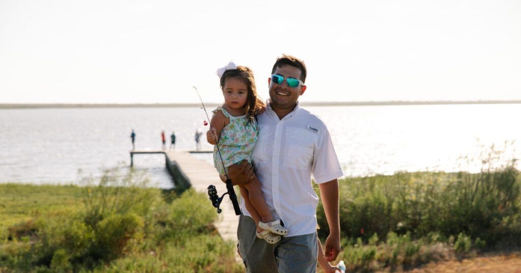 A man wearing sunglasses and a white shirt holds a young girl with a fishing pole, standing near a grassy shoreline with a dock and people fishing in the background.