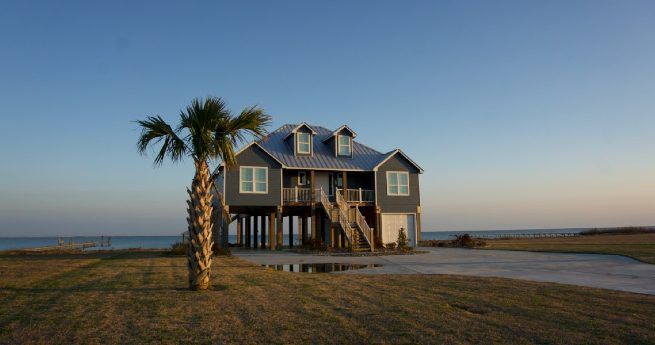 A beach house elevated on stilts with a metal roof and surrounded by open grass, with a single palm tree in the foreground and a clear sky overhead.