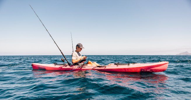 A person is sitting in a bright red fishing kayak on calm, open water, adjusting their fishing gear.