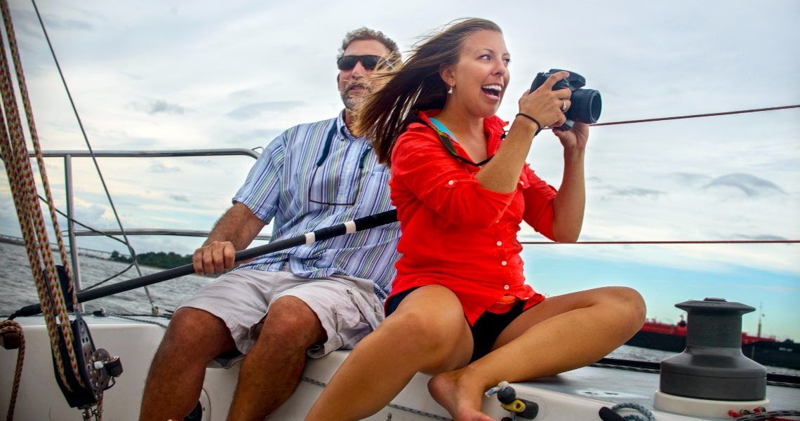 A woman enthusiastically takes photos with a camera while sitting on a sailboat, accompanied by a man steering.