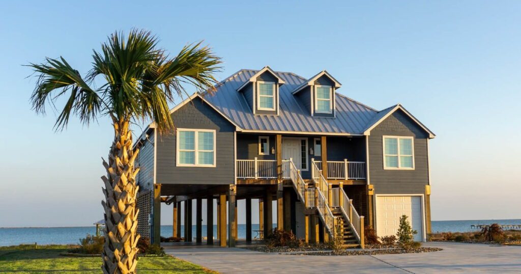 A modern stilt house painted dark blue stands prominently near the waterfront, surrounded by palm trees. The house has a large porch, multiple windows, and a tall staircase leading to the entrance. The water is visible in the background.