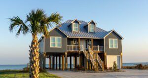 A modern stilt house painted dark blue stands prominently near the waterfront, surrounded by palm trees. The house has a large porch, multiple windows, and a tall staircase leading to the entrance. The water is visible in the background.