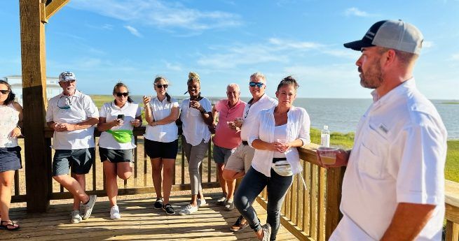 A group of casually dressed people stands on a wooden deck near the water, engaging in conversation and enjoying drinks. The weather is sunny with a bright blue sky.