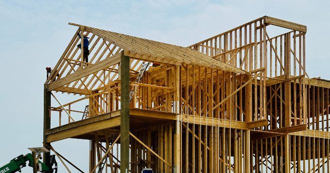 The wooden frame of a house under construction is shown in progress, with workers standing on the roof. The skeletal structure highlights the beams and supports of the multi-story build.