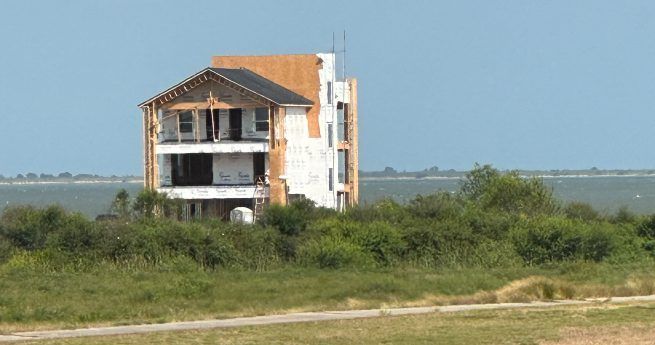 A three-story house in mid-construction stands on a grassy plot near the water, with exposed framework and insulation visible. The background shows water and a clear blue sky.