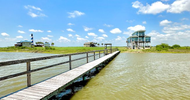 building a waterfront home in texas-houses and dock-4 A wooden dock stretches over calm water leading to a stilt house on the shoreline. The sky is clear with scattered clouds, and other houses are visible in the distance across the grassy land.