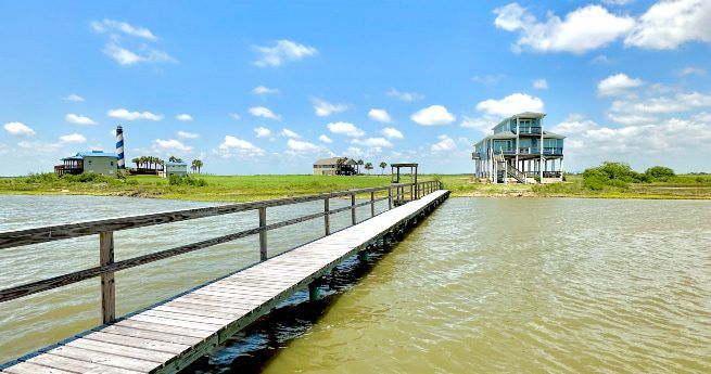 A wooden dock stretches over calm water leading to a stilt house on the shoreline. The sky is clear with scattered clouds, and other houses are visible in the distance across the grassy land.