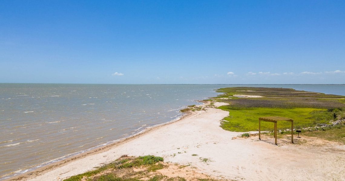 matagorda bay-coast-6 A stunning stretch of coastline with white sandy beaches meeting calm bay waters, surrounded by green vegetation under a bright blue sky.