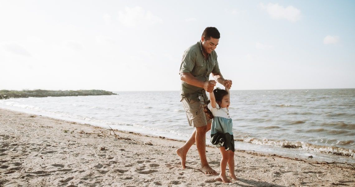 matagorda bay-father and daughter-5 A father joyfully playing with his young daughter along a sandy beach, lifting her by the arms as waves gently roll in the background.
