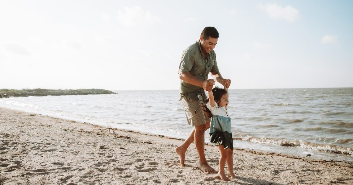 A father joyfully playing with his young daughter along a sandy beach, lifting her by the arms as waves gently roll in the background.