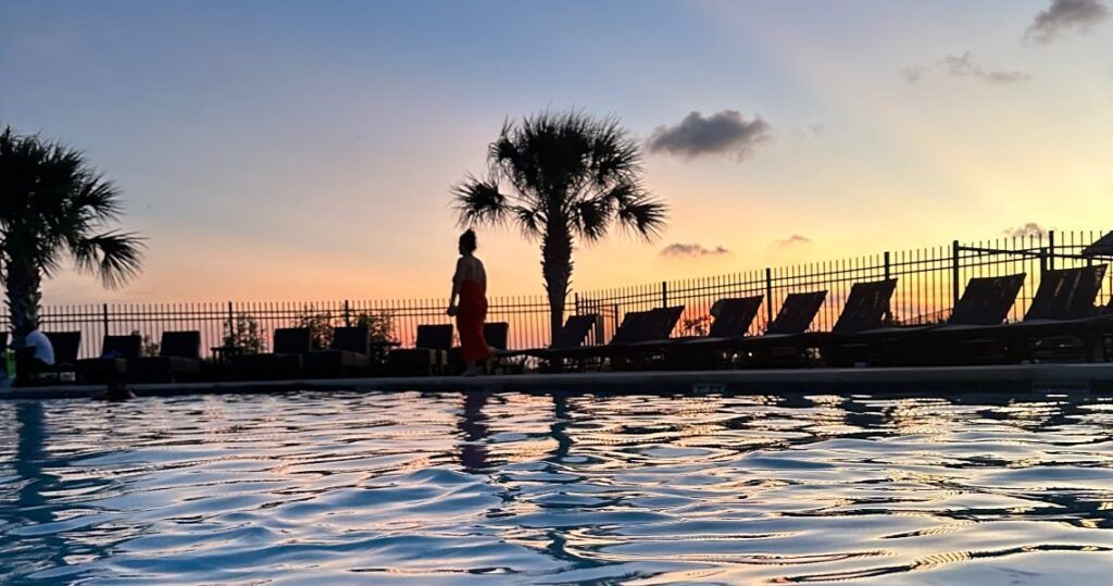 A serene poolside view at sunset, featuring a person walking along the edge of the pool with silhouettes of palm trees and lounge chairs in the background.