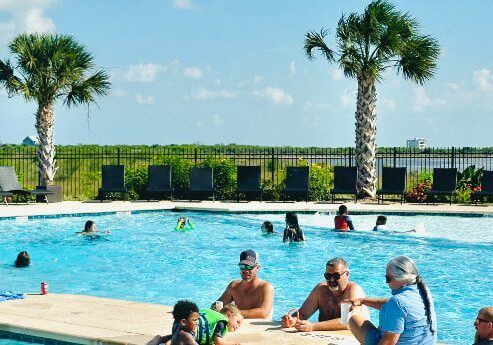 The image shows a group of people enjoying a sunny day at a swimming pool. Several adults and children are sitting or standing in the shallow end, socializing near the pool's edge. The pool area is surrounded by palm trees and a fence, with lounge chairs lined up in the background. Beyond the fence, lush greenery and a distant body of water create a scenic view.